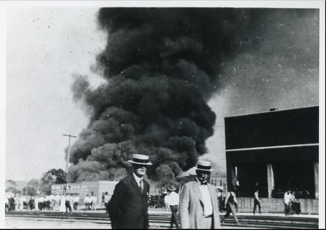 Two people stand near the railroad tracks across the street from a burning building during the 1921 Tulsa Race Massacre. The background shows a group of people standing and watching the building burn. - PHOTO COURTESY OF TULSA HISTORICAL SOCIETY & MUSEUM