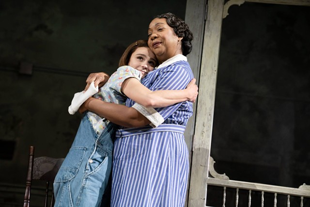 A young girl hugs an older woman, who wears a blue and white dress typically associated with household workers.